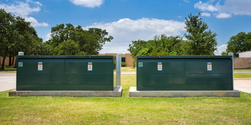 Two green underground distribution switchgear boxes on concrete pads. In the foreground is green grass. In the background are trees and a blue sky