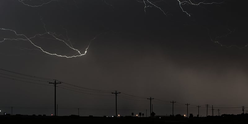 10 wooden poles on an overhead, 3-phase feeder at night in a rural area. Two large strokes of lightning and dark clouds line the sky above the feeder